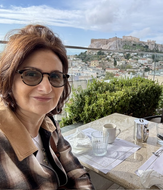 Stephanie looking at camera. She's seated at an outside table. Greece landscape in the background.